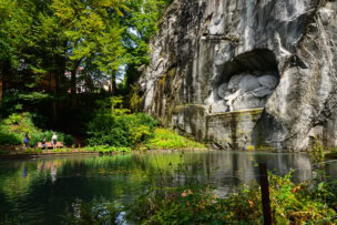 A rock face with a lion sculpture carved into it overlooks a pond, surrounded by lush greenery. Two people stand on the path beside the pond, enjoying one of the top things to do in Lucerne amidst this serene setting.
