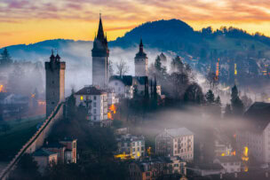 Foggy morning view of a medieval town reminiscent of Lucerne, with stone towers, scattered buildings, and trees set against rolling hills under a colorful sunrise sky. A perfect scene to inspire your travel tips and things to do for an unforgettable journey.