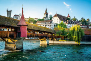Explore the rustic wooden bridge in Lucerne, featuring a charming red spire as it crosses a gentle river. With historic buildings and a church tower in the backdrop against a clear blue sky, it's one of the top things to do for any travel enthusiast.