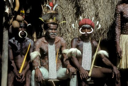 Three men in traditional attire and body paint sit in front of a thatched structure, wearing headdresses, necklaces, and facial piercings. Two hold yellow spears, one wears a red headband. A traveler nearby listens attentively to their guide explaining the rich cultural heritage on this fascinating tour.