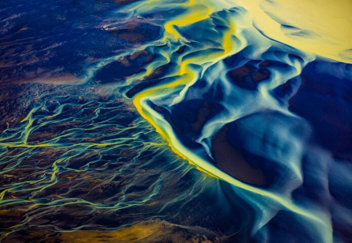 Aerial view of braided river channels and sediment patterns with vibrant blue and yellow hues, surrounded by dark land.