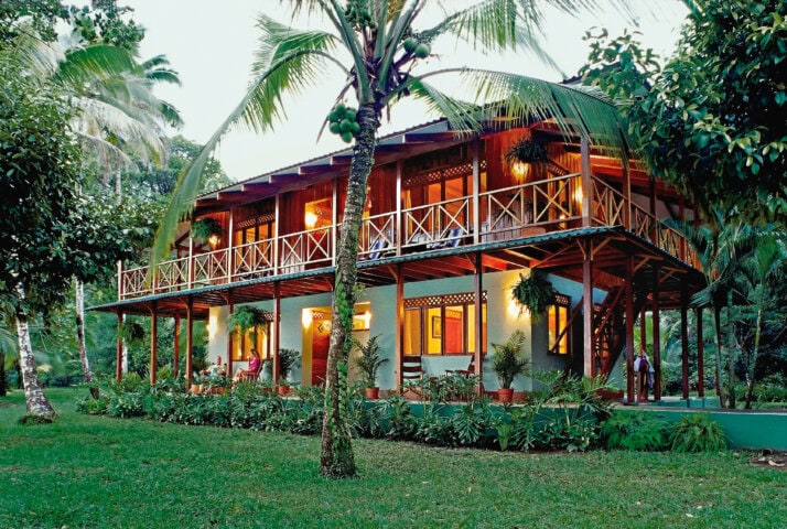 A two-story house with wooden balconies in Costa Rica, surrounded by lush greenery and palm trees, illuminated by warm lights at dusk.
