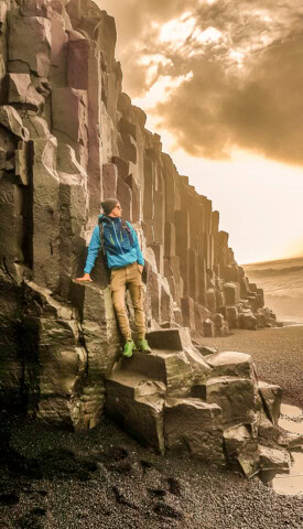 A person in outdoor gear leans against tall basalt columns at a rocky beach in Iceland, under a dramatic sky.