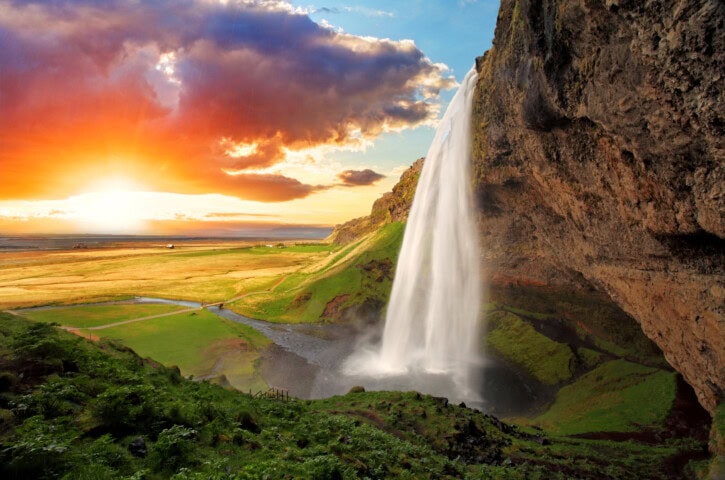 A waterfall cascades from a rocky cliff into a grassy valley, with the sun setting and colorful clouds in the sky, creating an idyllic scene perfect for travel enthusiasts exploring Iceland's natural wonders.
