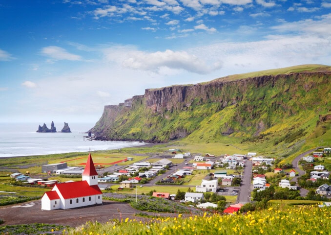 A small Icelandic village with a red-roofed church is situated on a grassy landscape by the sea, with tall cliffs and coastal rock formations in the background under a partly cloudy sky—a perfect spot for travel and tourism enthusiasts.