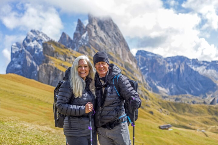 A smiling couple in hiking gear stands on a grassy hill with hiking poles, set against a backdrop of jagged mountains and cloudy skies.