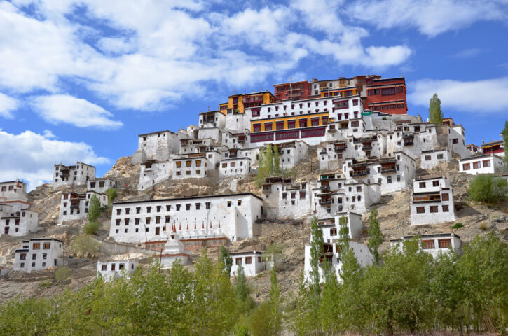 A large, multi-tiered white monastery with red and yellow accents sits on a hillside under a blue sky with scattered clouds, surrounded by greenery. This serene sanctuary evokes the mythical Shangri-La, beckoning travelers to embark on a journey of discovery and tranquility.