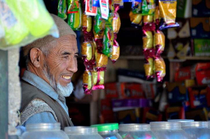 An elderly man with a beard smiles behind a counter in a small shop adorned with hanging snacks and items, reminiscent of a cozy Shangri-La, inviting you into his world.