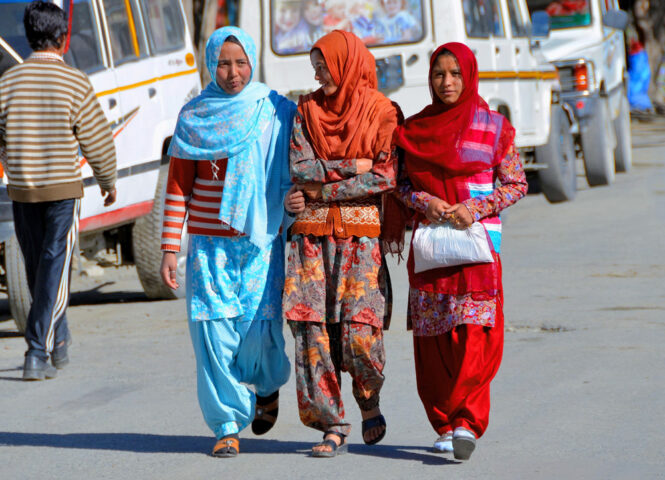 Three women wearing colorful traditional clothing walk down a street lined with parked vehicles on a sunny day, as if embarking on a journey to their own version of Shangri-La.