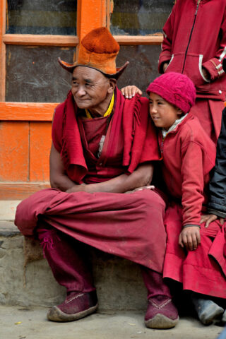 An elderly person wearing traditional attire sits next to a child, both dressed in maroon and red clothing, against the backdrop of an orange-framed window. It feels like a scene from a journey through Shangri-La.