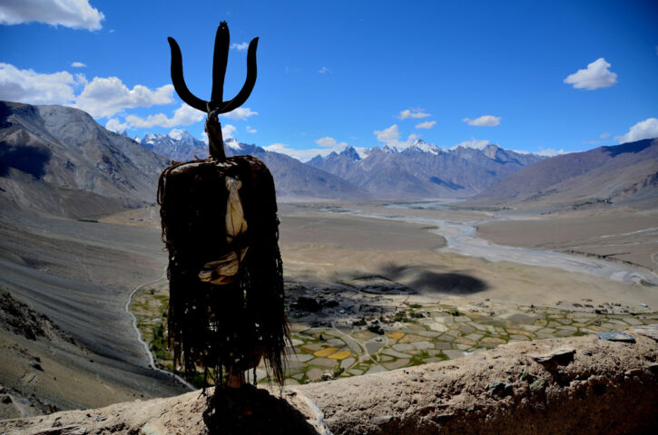A tribal statue with a trident on its head overlooks a vast, mountainous landscape reminiscent of Shangri-La, with a river winding through a valley and a cluster of small settlements below. The sky is clear and blue.