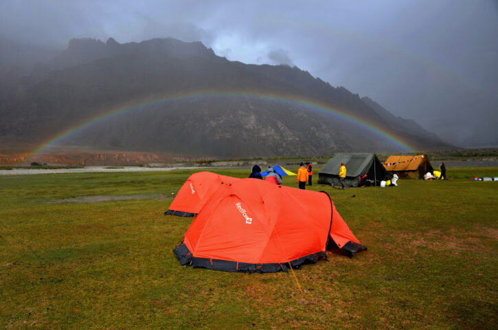 Red and green tents set up on a grassy field with people standing nearby, a rainbow arcs over the mountain range in the background under a cloudy sky, creating a scene reminiscent of a Journey to Shangri-La.