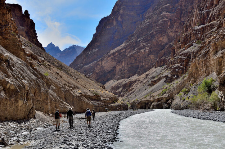A group of hikers embarks on a journey along a rocky path beside a flowing river in a steep, rugged canyon under a partly cloudy sky, reminiscent of an earthly Shangri-La.