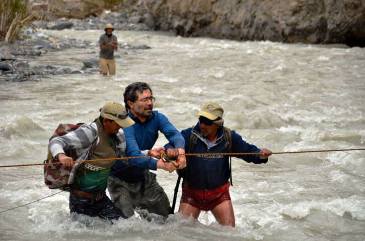 Three men are wading through a fast-moving river, holding onto a rope for support, as part of their journey to Shangri-La, with a fourth man standing on the rocky shore in the background.