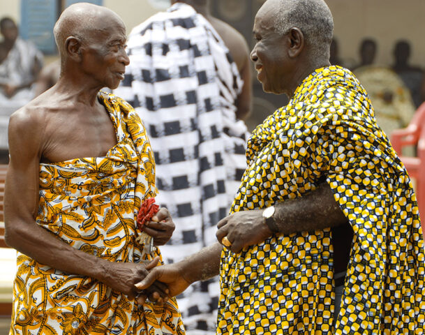Two elderly men in brightly patterned traditional clothing are shaking hands and conversing outdoors on a sunny day, reminiscent of a scene from "The Ashanti King and I.