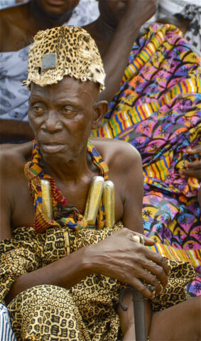 An elderly Ashanti King wearing a traditional patterned robe and leopard print hat sits surrounded by people, holding a decorative staff, and adorned with a necklace featuring large cylindrical beads.