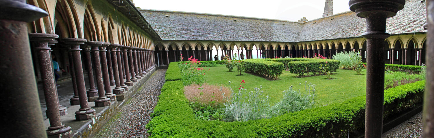 Exploring the medieval cloister at Mont Saint Michel, you'll find arched columns surrounding a rectangular garden adorned with neatly trimmed hedges and scattered red flowers.