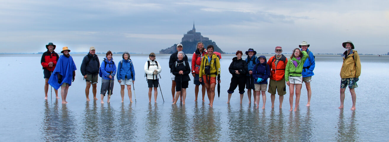 A group of people stand barefoot in shallow water, dressed for hiking. Mont Saint-Michel, the Wonder of the West, is visible in the background under a cloudy sky.
