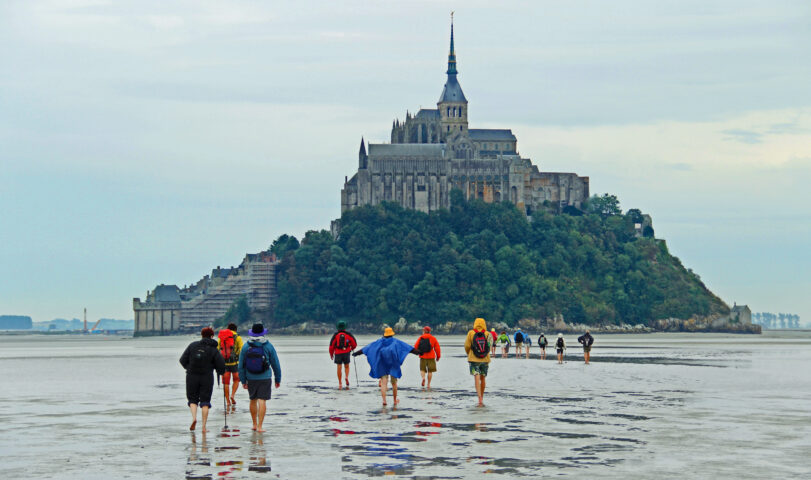 A group of people walking towards Mont Saint-Michel, the Wonder of the West, a historic monastery located on an island in Normandy, France, during low tide.
