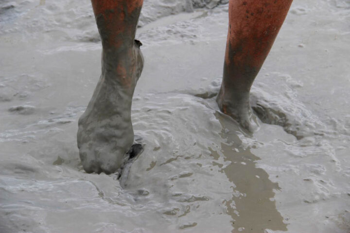 Close-up of a person's legs and feet partially submerged in thick, gray mud—a true Wonder of the West. The legs are covered in mud up to the knees.