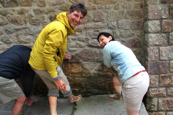 Three people, two in light-colored shorts and jackets, and one in dark shorts, are playfully washing their feet with water from a stone wall spout. The person in the yellow jacket, showcasing the simple joys of the Wonder of the West, is smiling at the camera.