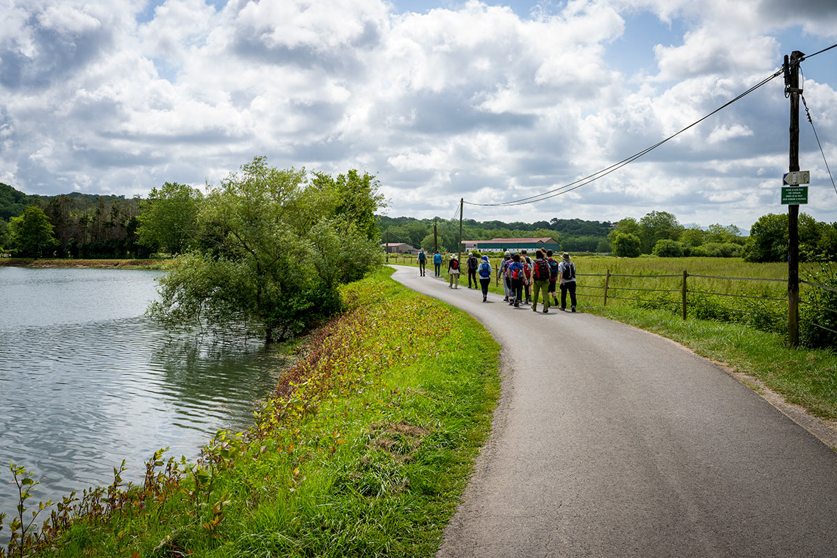 A group of people with backpacks hike along a paved path next to a body of water and green fields in the Basque Country under a cloudy sky.