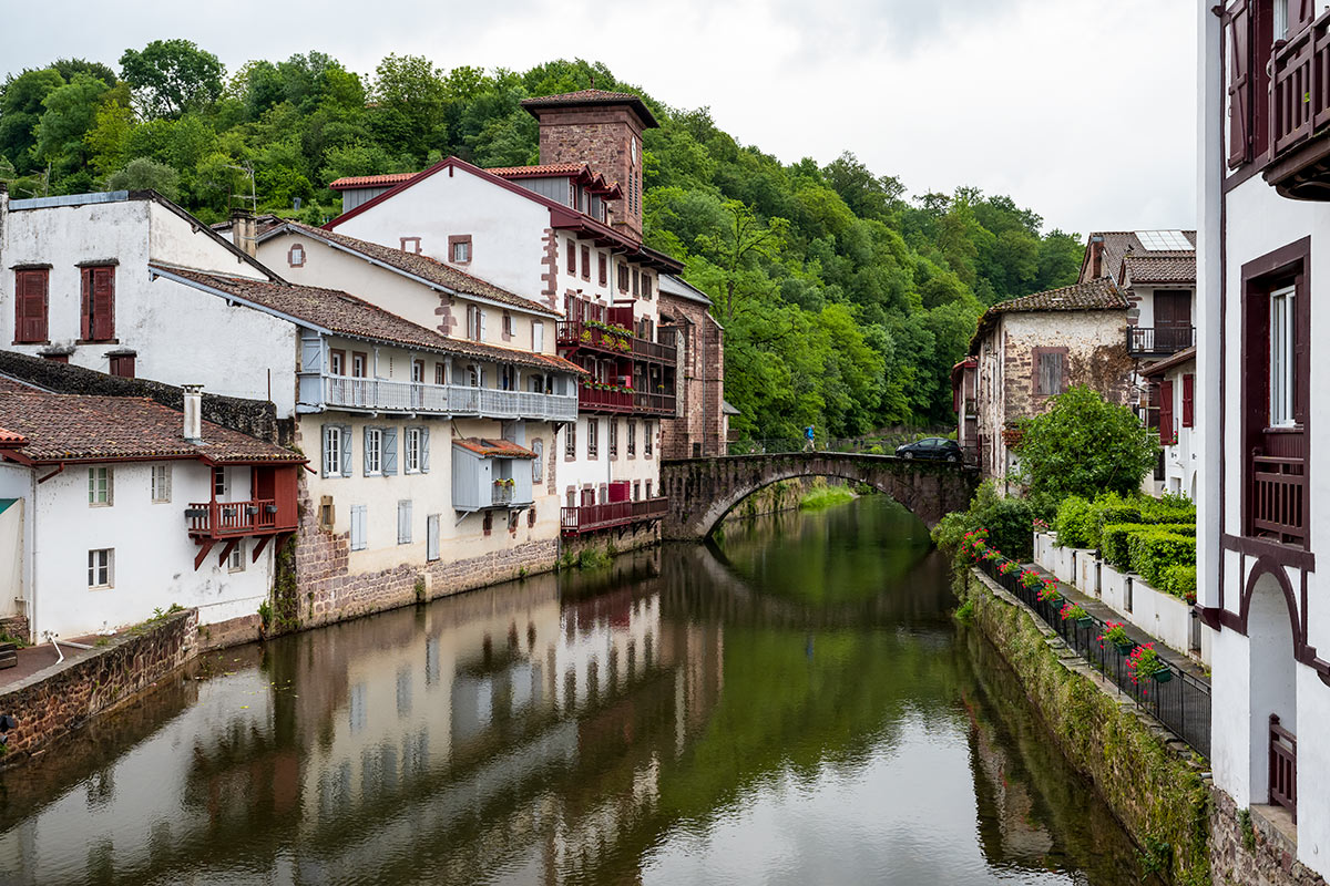 A narrow river flows through a quaint village with white and brick buildings and a stone bridge, surrounded by lush greenery, perfect for hiking enthusiasts exploring the serene Basque Country.