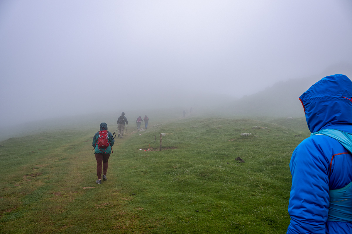 Hikers in colorful jackets walk along a foggy grassy trail in the Basque Country, with limited visibility due to dense mist.