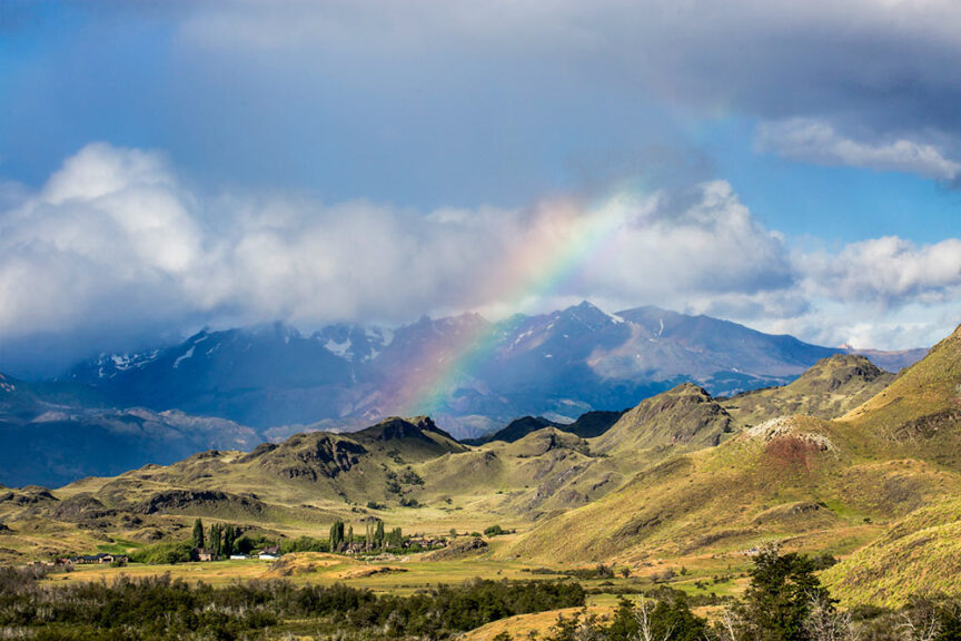 A scenic view of a mountainous landscape in the Patagonian National Parks with a rainbow arching over the hills under a partly cloudy sky. The vast beauty of these 11 million acres in Chile is truly breathtaking.
