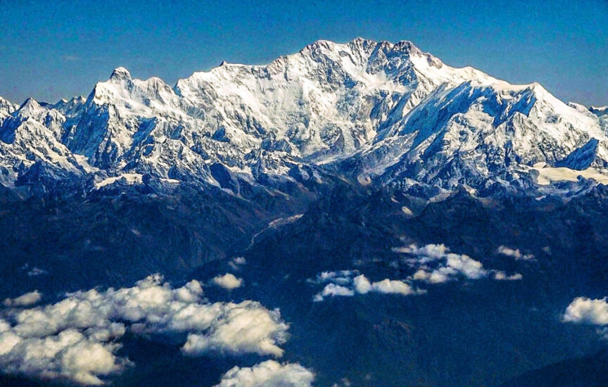 A snow-covered mountain range stretches above the clouds under a clear blue sky, with scattered clouds in the foreground. The terrain transitions from snowy peaks to rocky lower slopes.