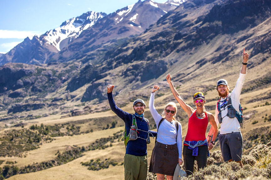 Four hikers, three of whom are raising their arms, stand on a grassy field with a mountainous landscape and snowy peaks in the background on a sunny day in one of Chile's Patagonian National Parks.