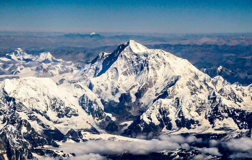 Aerial view of a snow-covered mountain peak surrounded by other snowy mountains under a clear blue sky, inviting travelers to embark on an unforgettable adventure above the clouds.