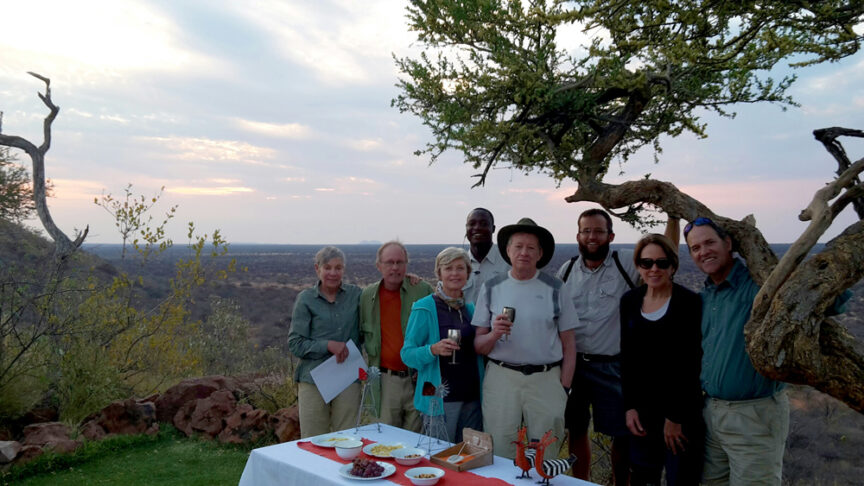 A group of eight people stand outdoors around a table with food and drinks, with a scenic landscape featuring a tree and distant hills in the background. They appear as if they're enjoying a feast fit for the Kings of Africa.