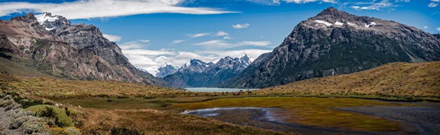 A vast mountainous landscape with snow-capped peaks, blue sky, and a serene lake in the distance. Grassy plains in the foreground with patches of greenery create the perfect setting for outdoor clothing enthusiasts or those seeking adventure gear for their next Patagonia expedition.