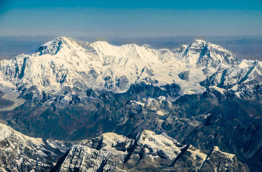 Snow-covered mountain range with prominent peaks reaching above the clouds under a clear blue sky. The lower elevations have less snow and more rocky terrain.