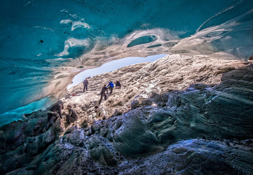 People wearing Patagonia outdoor clothing explore a rocky terrain beneath an arch of ice, with daylight filtering through the blue-tinted ice above.