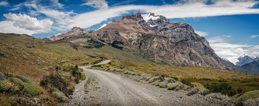 A winding dirt road leads through a mountainous landscape with rocky peaks and sparse vegetation under a partly cloudy blue sky, perfect for testing outdoor clothing and adventure gear in the rugged wilderness of Patagonia.