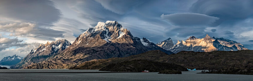 Panoramic view of a mountain range with snow-capped peaks, dramatic clouds, and a body of water in the foreground. A small boat is visible on the water, evoking the remote beauty of Patagonia.