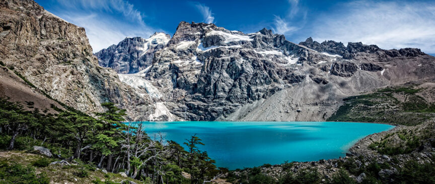 A bright turquoise lake sits at the base of snow-capped rocky mountains in Patagonia under a clear blue sky, with dense green trees in the foreground and adventurers clad in sustainable outdoor clothing exploring the terrain.