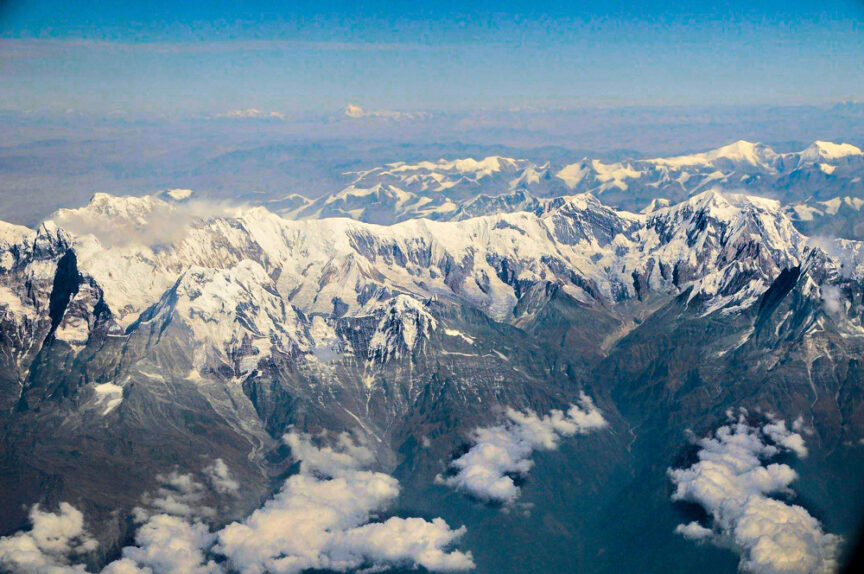 Aerial view of a mountain range with snow-covered peaks, scattered clouds below, all under a clear sky—truly a scene from above the clouds.
