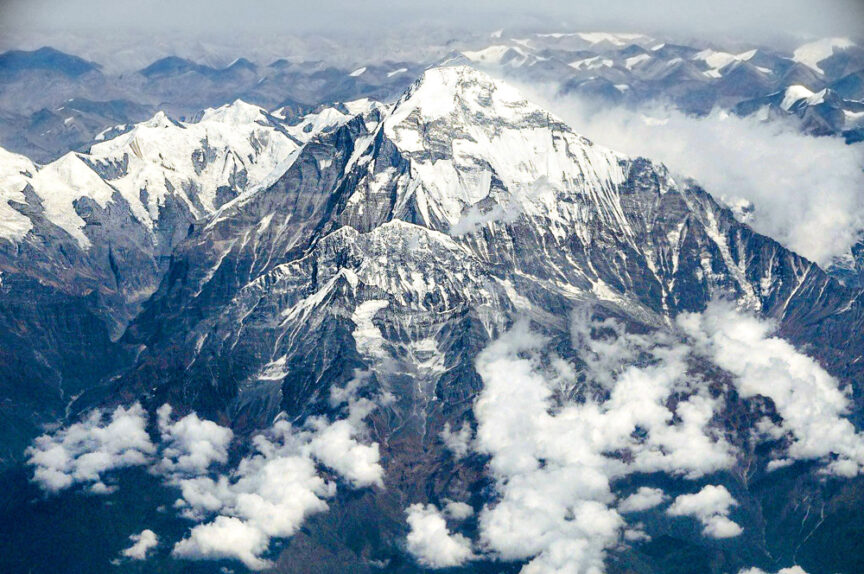 Aerial view of snow-covered mountain peaks with clouds hovering at lower elevations. The largest peak, rising boldly above the clouds, is surrounded by other rugged, smaller peaks.