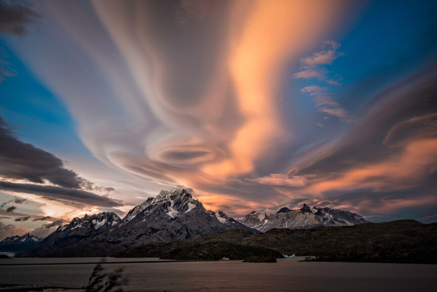 Dramatic cloud formations above rugged, snow-capped mountains in Patagonia at sunset, with a calm body of water in the foreground—a perfect setting for testing outdoor clothing and adventure gear.