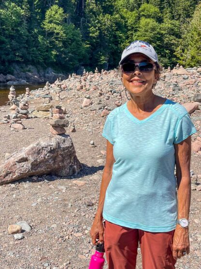 A woman wearing a light blue shirt, sunglasses, and a cap stands on a rocky beach at Canada's Bay of Fundy, holding a water bottle with stacked stone formations in the background, ready for her adventures.