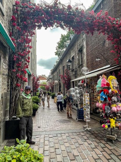 A narrow cobblestone street lined with shops and vendors, featuring a man in a green jacket standing under an archway adorned with red flowers. People are walking and browsing nearby, reminiscent of picturesque adventures often found near the Bay of Fundy in Canada.