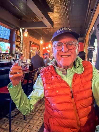 A smiling person wearing a hat, glasses, and an orange vest holds a drink while reminiscing about their adventures in the Bay of Fundy, sitting in a bar with a lit ambiance.
