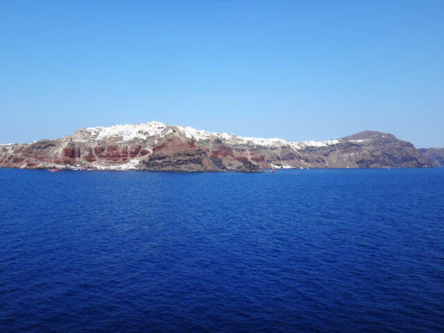 A view of a rocky island with white buildings on top, surrounded by the deep blue Aegean Sea under a clear sky, showcasing vibrant colors.