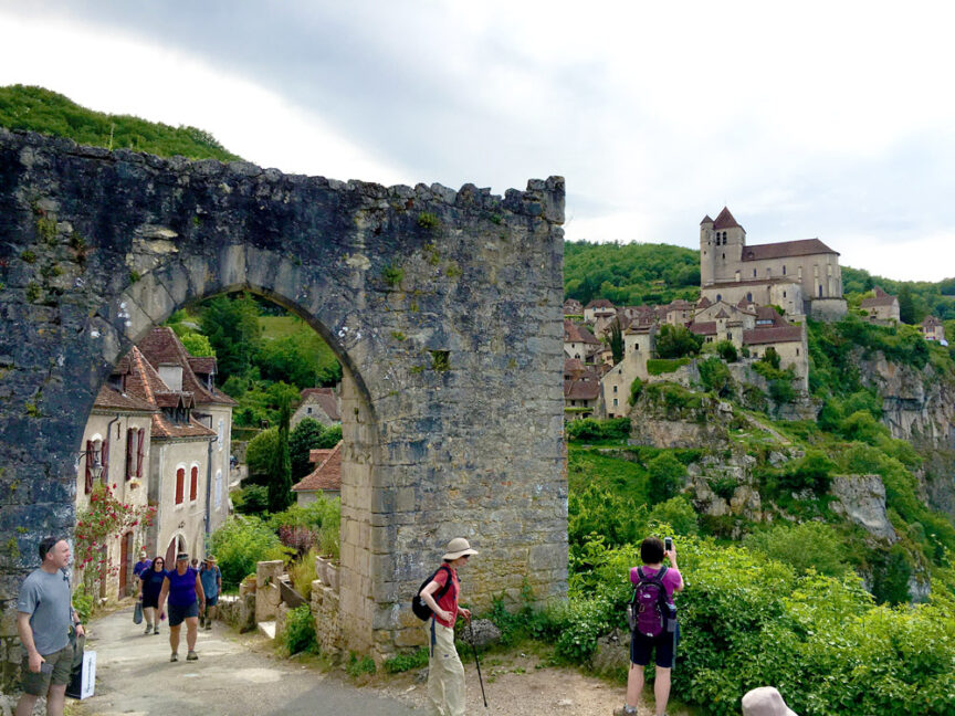 Tourists walking through a stone archway towards the delightful hillside village in Lot Valley, with a charming church and buildings nestled among greenery, under a cloudy sky.