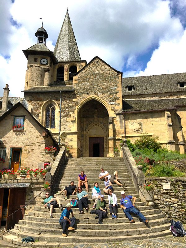 A group of people delight in sitting on the steps of an old stone church with a clock tower, surrounded by flowers and greenery in the enchanting Lot Valley, under a partly cloudy sky.