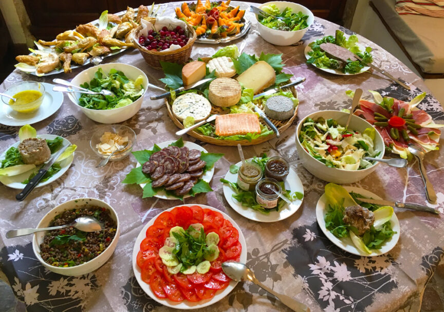 An assortment of delightful dishes, including salads, cheeses, meats, sliced tomatoes with cucumbers, and condiments, arranged on a round table covered with a patterned tablecloth inspired by the charm of Lot Valley.