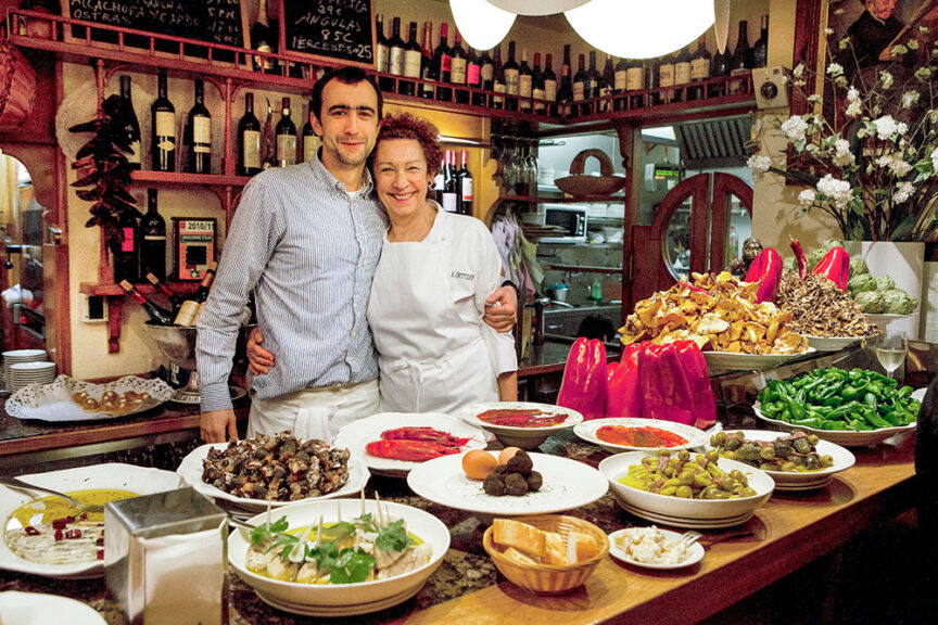 A man and a woman, resembling seasoned travelers, stand smiling behind a European-style restaurant counter with various dishes and ingredients, including fresh vegetables and seafood, displayed in front of them.